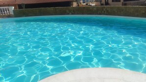 En pool med klart vatten och tydliga vågmönster, omgiven av en betongmur och en byggnad med ett tak täckt av tegelpannor i bakgrunden.