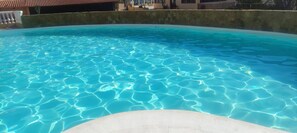 Ein klares Schwimmbecken mit erkennbaren Wassermustern, umgeben von einer Betonmauer und einem Gebäude mit einem Ziegeldach im Hintergrund.