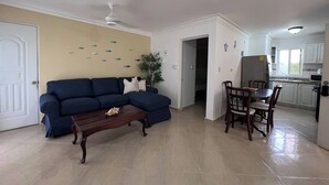A living room with a sofa, coffee table, and a dining area with chairs and a table. There is a ceiling fan, a plant, and a kitchen visible in the background. - Garden Condos 48 - Friendly 1BR APT, Sosua Center! (Sosúa)
