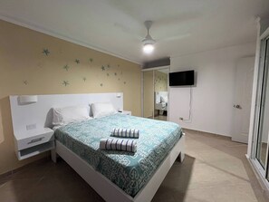 A bedroom with a bed, nightstands, a ceiling fan, a TV mounted on the wall, and a bathroom visible through an open door. - Garden Condos 48 - Friendly 1BR APT, Sosua Center! (Sosúa)