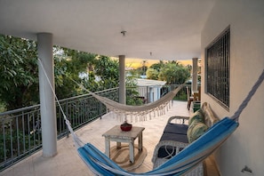 A balcony with a hammock, a small table with a plant, a sofa with cushions, and a view of greenery and a building. - COZY 7BR Villa  BIG POOL  Close to beach! (Sosúa)