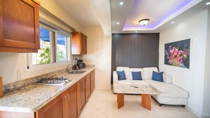 A modern kitchen with wooden cabinets, a gas stove, and a countertop. There is a white sofa with blue cushions and a coffee table in the living area. A painting of coral is on the wall. - CDC 2 Studio with Bar, Walkable to Beach & Fun (Sosúa)