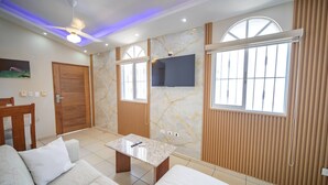 Un salon moderne avec un canapé, une table basse, un téléviseur et une grande fenêtre équipée de stores.