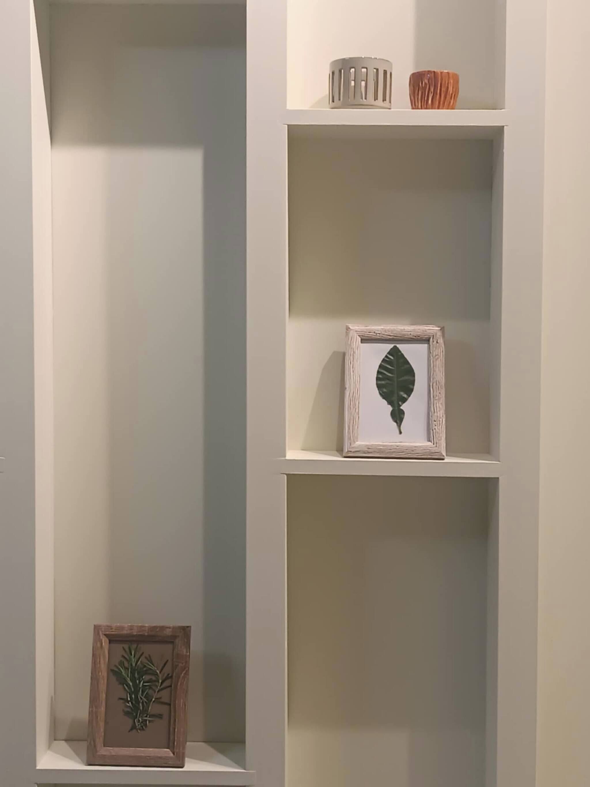 A white shelving unit with a framed leaf, a small framed plant, and two decorative objects on the top shelf.