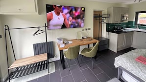 Habitación compacta con televisor, una pequeña mesa de comedor, dos sillas, una banca, radiador, zona de cocina con armarios y una ventana.
