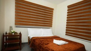 A hotel room with a bed, a bedside table with a water bottle and a plate of fruit, and a window with blinds. - Kodai Blossom Cottages (Kodaikanal)
