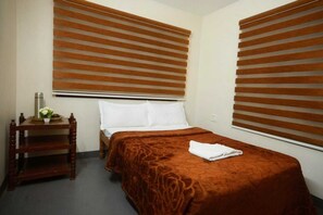 A hotel room with a bed, a bedside table with a water bottle and a plate of fruit, and a window with blinds. - Kodai Blossom Cottages (Kodaikanal)