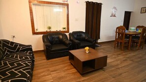 A living room with a sofa, two armchairs, a coffee table, and a dining area with a table and chairs. - Kodai Blossom Cottages (Kodaikanal)