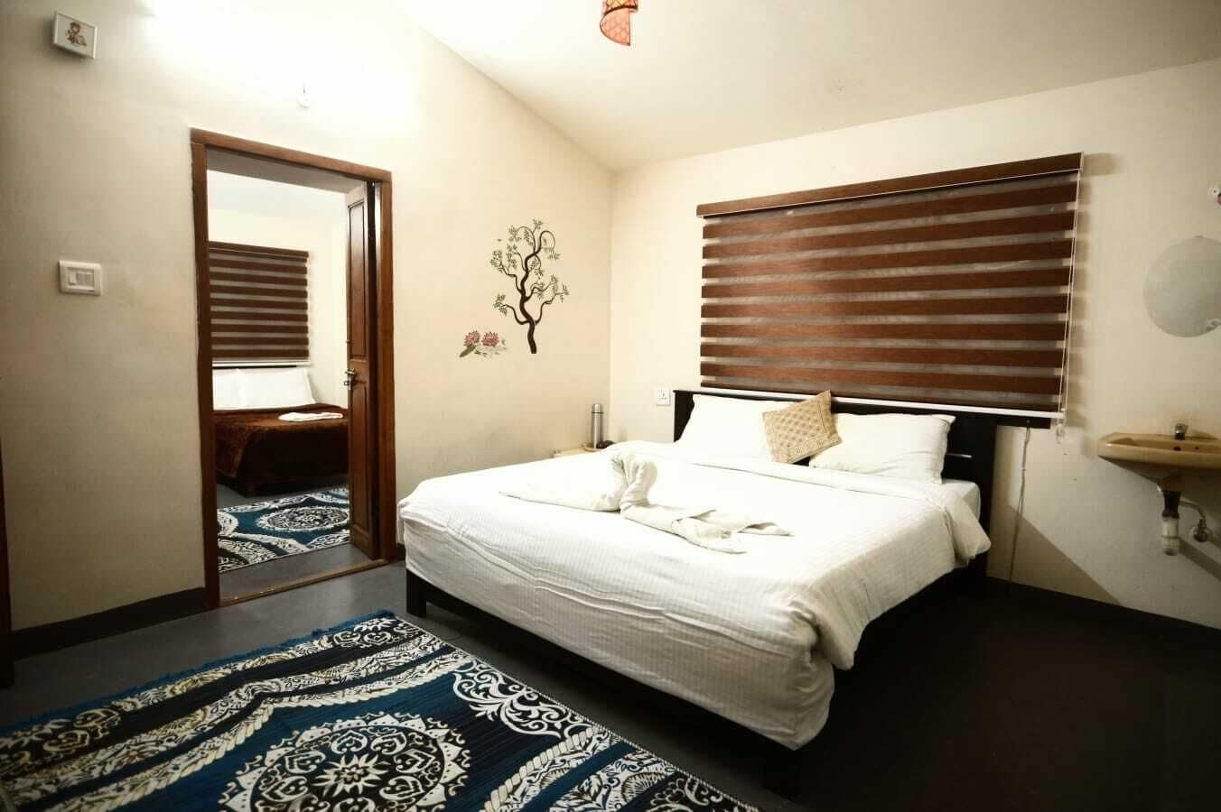 Un dormitorio con cama, una mesita de noche, estante en la pared, ventana con persianas, puerta y una alfombra estampada.