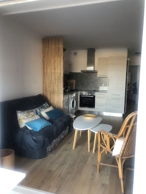 Ein gemütlicher, kompakter Wohnraum mit einer Couch, einem kleinen Tisch, Korbstühlen, einer Waschmaschine und einer Küchenzeile.