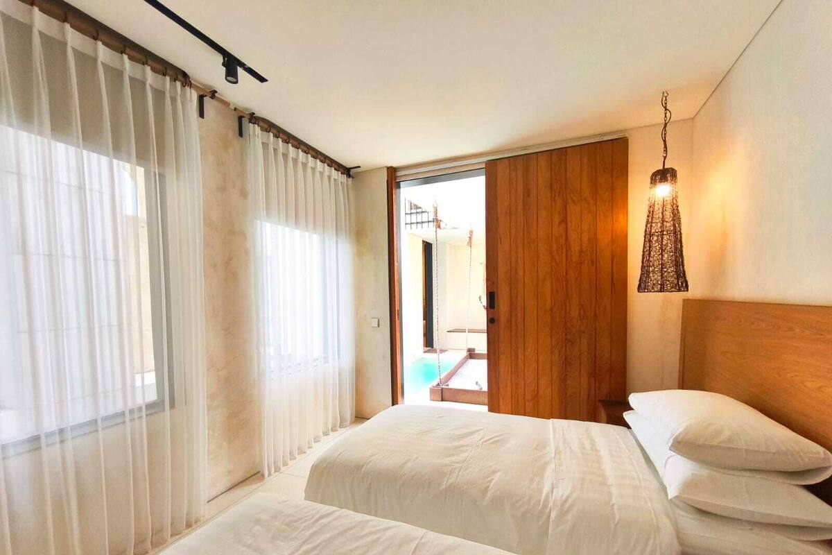 Une chambre d’hôtel avec deux lits, une tête de lit en bois, un luminaire suspendu et une vue sur une piscine extérieure.