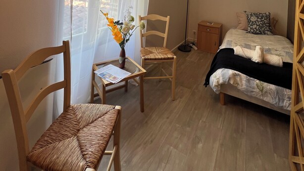 Un dormitorio con cama, dos sillas de madera, una mesita con un florero y una lámpara de pie.