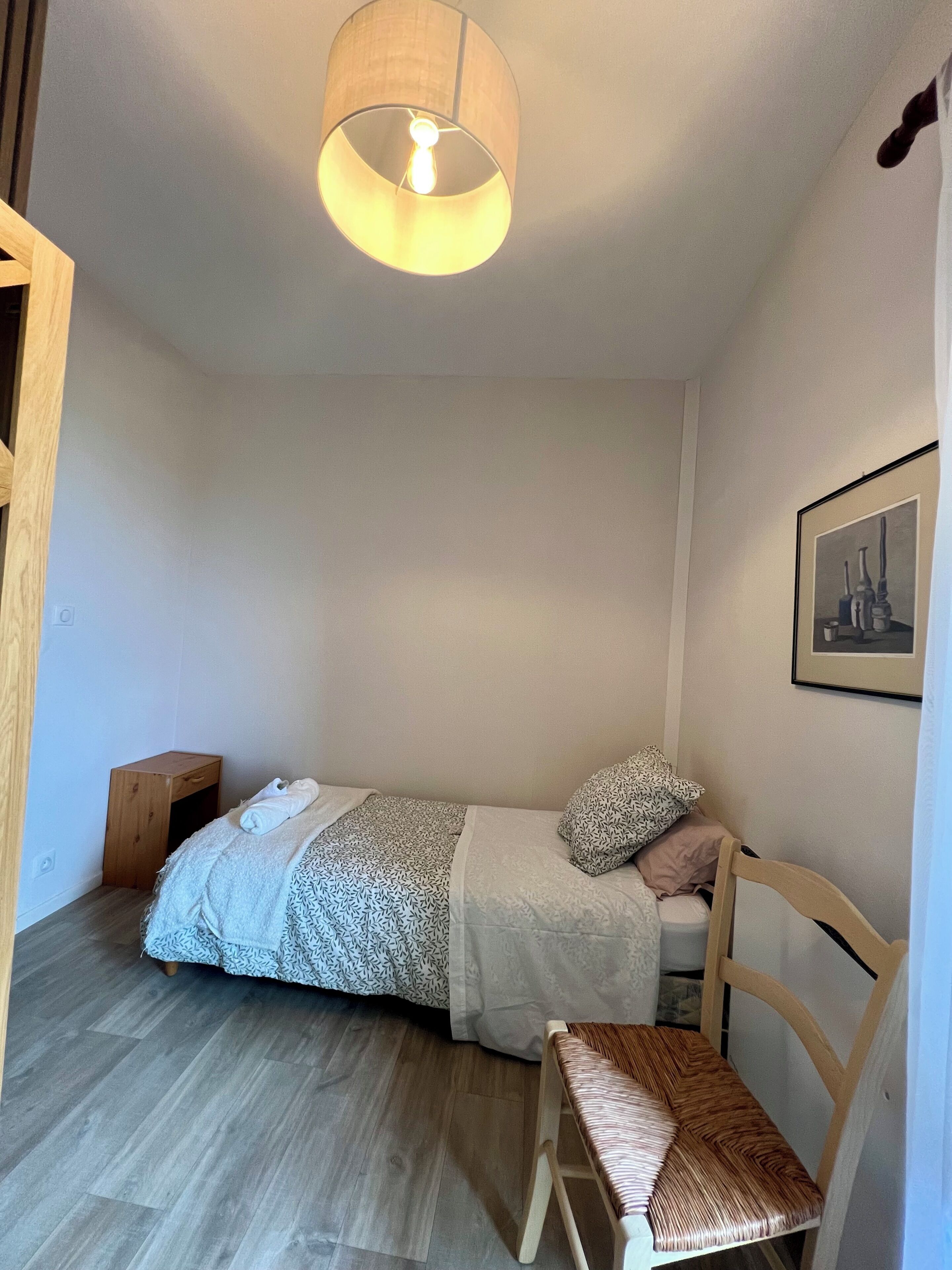 Un dormitorio pequeño y ordenado con una cama individual, una mesita de noche de madera, una silla y una lámpara colgante.