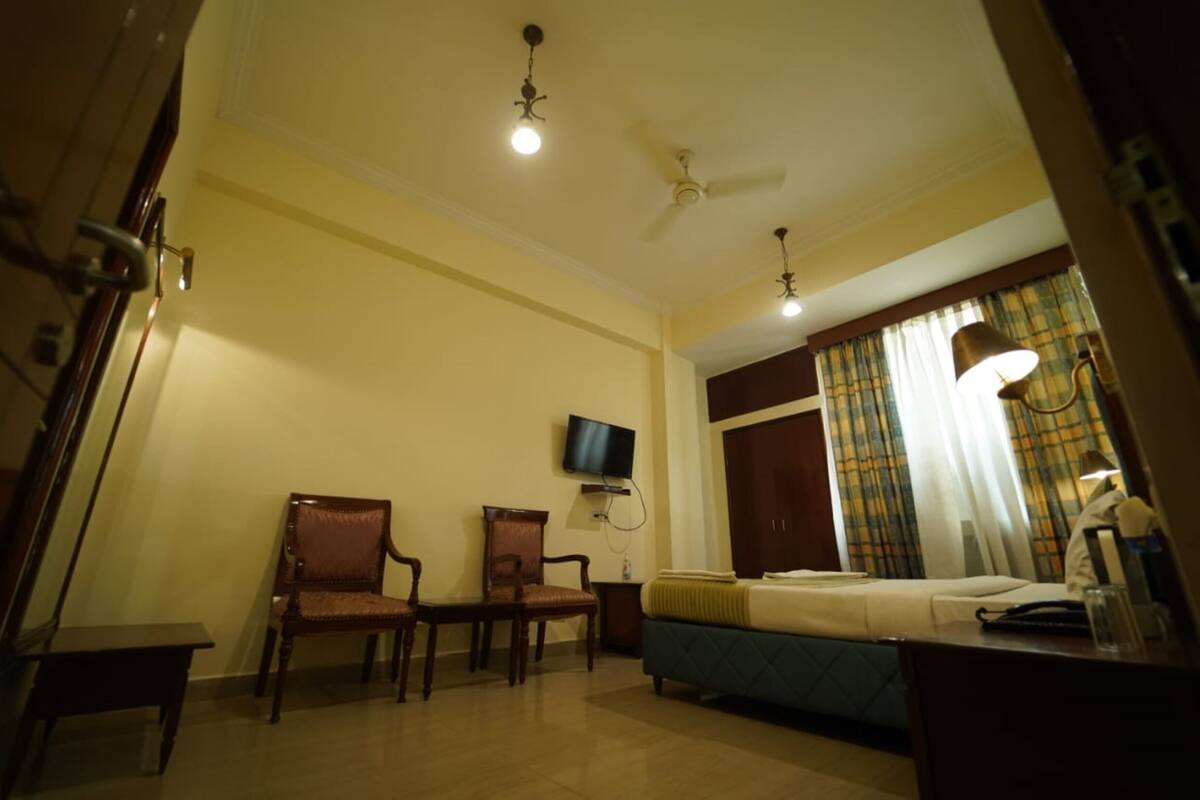 Habitación de hotel con cama, dos sillas, televisor, ventilador de techo y cortinas.