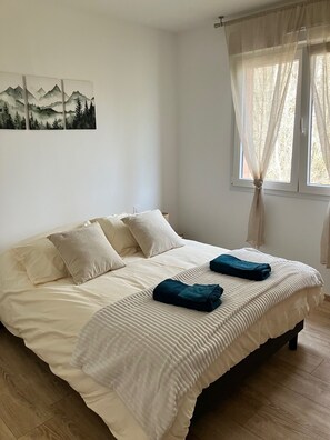 A neatly made bed with pillows and a blanket, a window with sheer curtains, and a wall-mounted painting of a mountain landscape. - Cosy house close to nature, 6/8 people, 10 min from FRAISPERTUIS park (La Bourgonce)