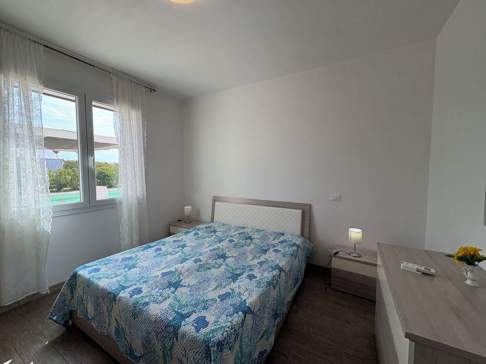 Een slaapkamer met een bed, nachtkastje, lamp en een raam met gordijnen.