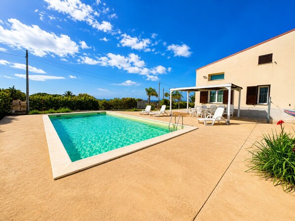 Una piscina rettangolare con acqua cristallina, circondata da un patio sabbioso. Sono presenti sedie a sdraio e una zona relax coperta con vista sull'oceano.