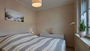 Un dormitorio con cama, lámparas de noche, mesita de noche, cómoda, un cuadro y una ventana con una planta.