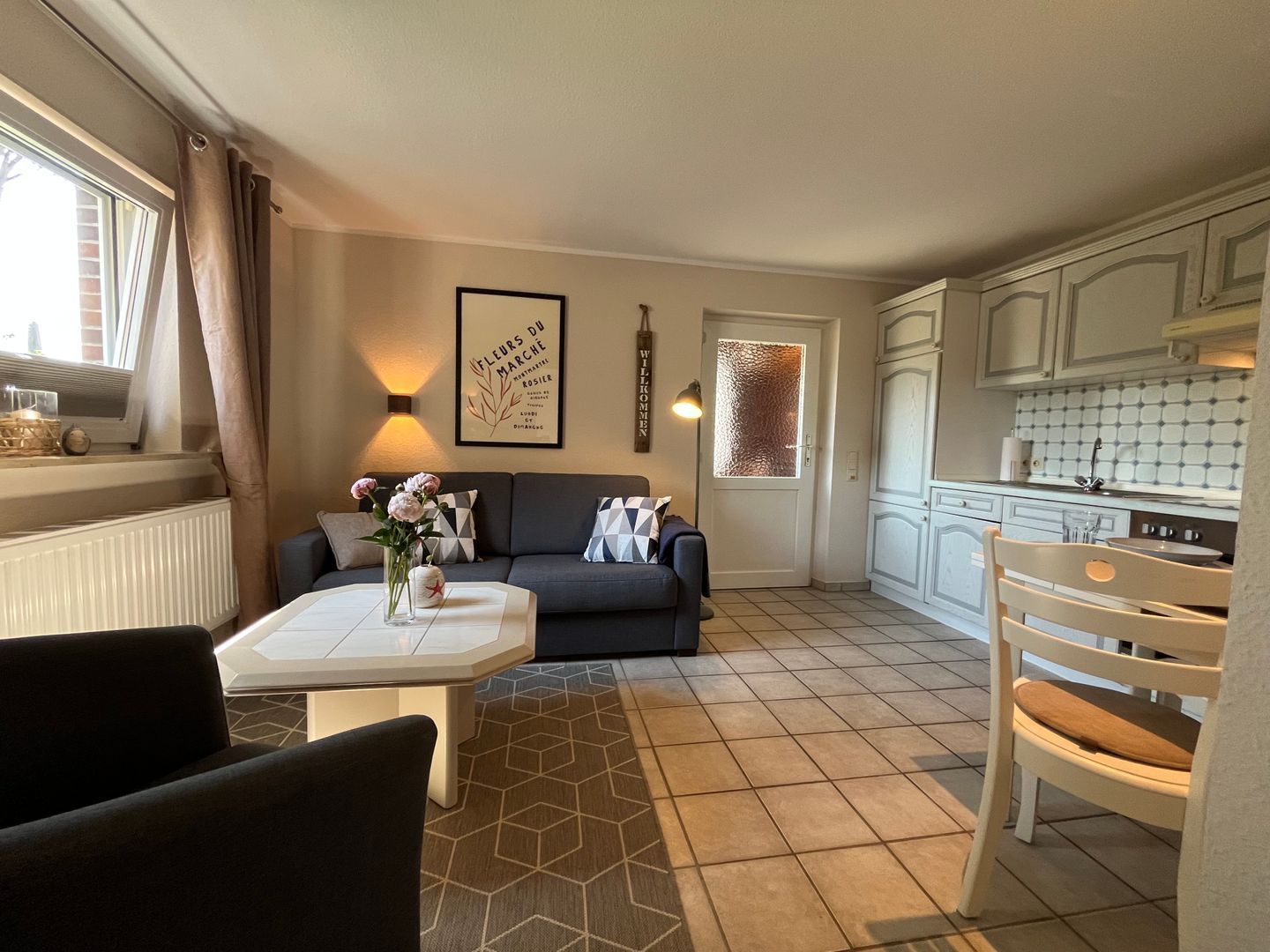 Una sala de estar con un sofá, una mesa de centro con flores, una cocina con gabinetes blancos y una puerta con vidrio esmerilado.