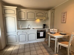 A kitchen with white cabinetry, a tiled backsplash, a stove, a sink, and a dining table with chairs. - Separate bungalow, 30sqm for up to 2 people - Nordgang 6 (Sylt-Ost)