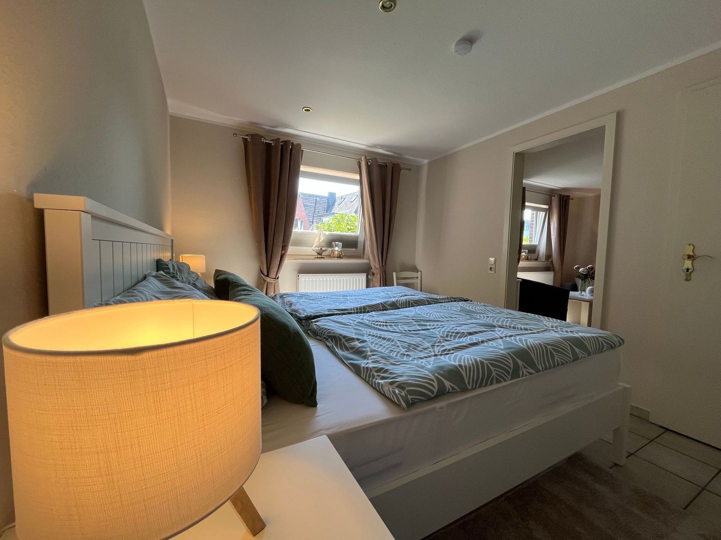 A bedroom with a bed, bedside table, lamp, and a view of the outside.