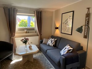 A living room with a sofa, a small table with a vase of flowers, a window with curtains, a wall-mounted lamp, and a framed welcome sign. - Separate bungalow, 30sqm for up to 2 people - Nordgang 6 (Sylt-Ost)