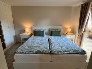 A double bed with leaf-patterned bedding, two bedside tables with lamps, a wardrobe, and a window with curtains. - Detached bungalow, 30 m² for up to 2 people - Nordgang 6 (Sylt-Ost)