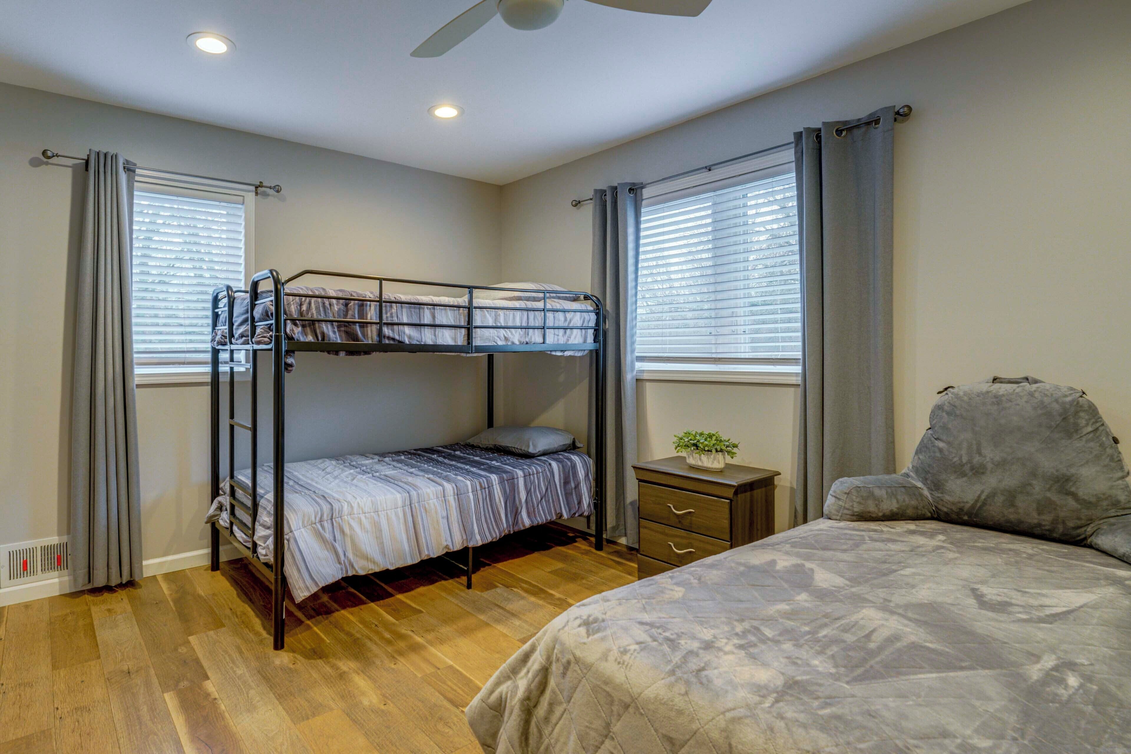A room with a bunk bed, a single bed, a small bedside table with a plant, and a ceiling fan.