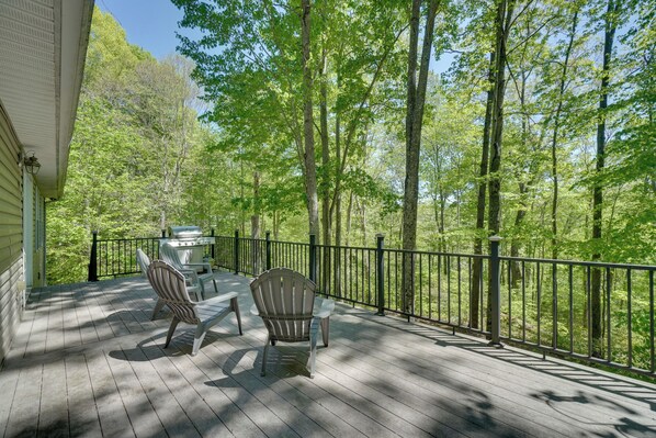 Eine Holzterrasse mit Adirondack-Stühlen, einem Grill und Blick auf einen Wald.