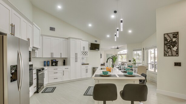 A modern kitchen with white cabinets, a central island with bar stools, and a dining area with a view of the outdoors. - Waterfront Pool Home by Park Pickleball Sleeps 10 (Cape Coral)