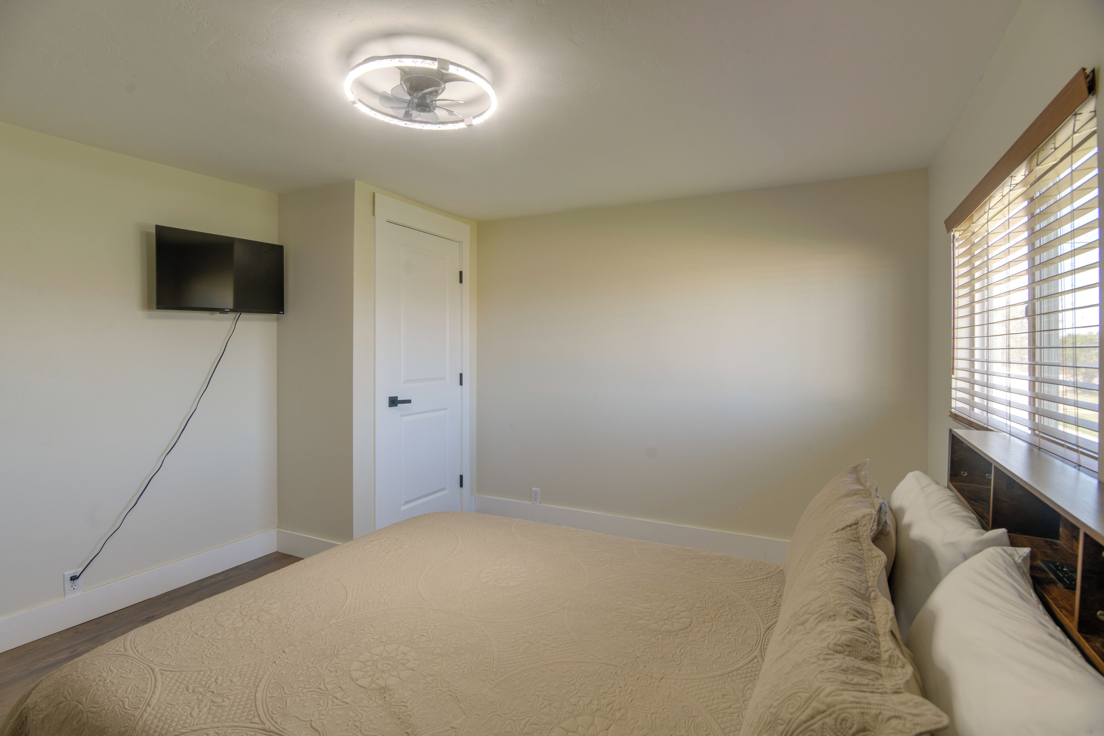 A bedroom with a bed, a television mounted on the wall, a door, and a window with blinds.