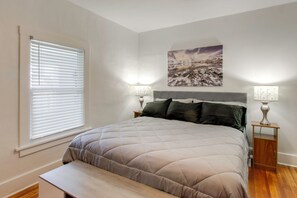 Un dormitorio con cama, lámparas de noche, mesita de noche, un banco, una ventana con persianas y un cuadro colgado en la pared.