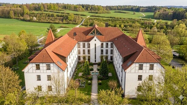 Een groot, symmetrisch gebouw met een centrale binnenplaats, omgeven door weelderige begroeiing en een pittoresk landschap.