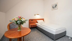 Een eenpersoonsbed met een houten hoofdbord, een nachtkastje met een lamp, een klein houten nachtkastje, een stoel en een vaas met bloemen.