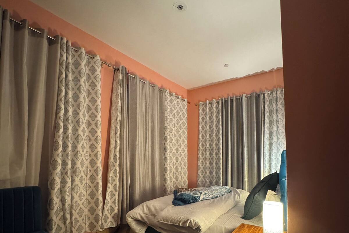 Une chambre à coucher avec un lit, une table de chevet avec une lampe, une chaise bleue, des rideaux à motifs et un tapis à motifs géométriques.
