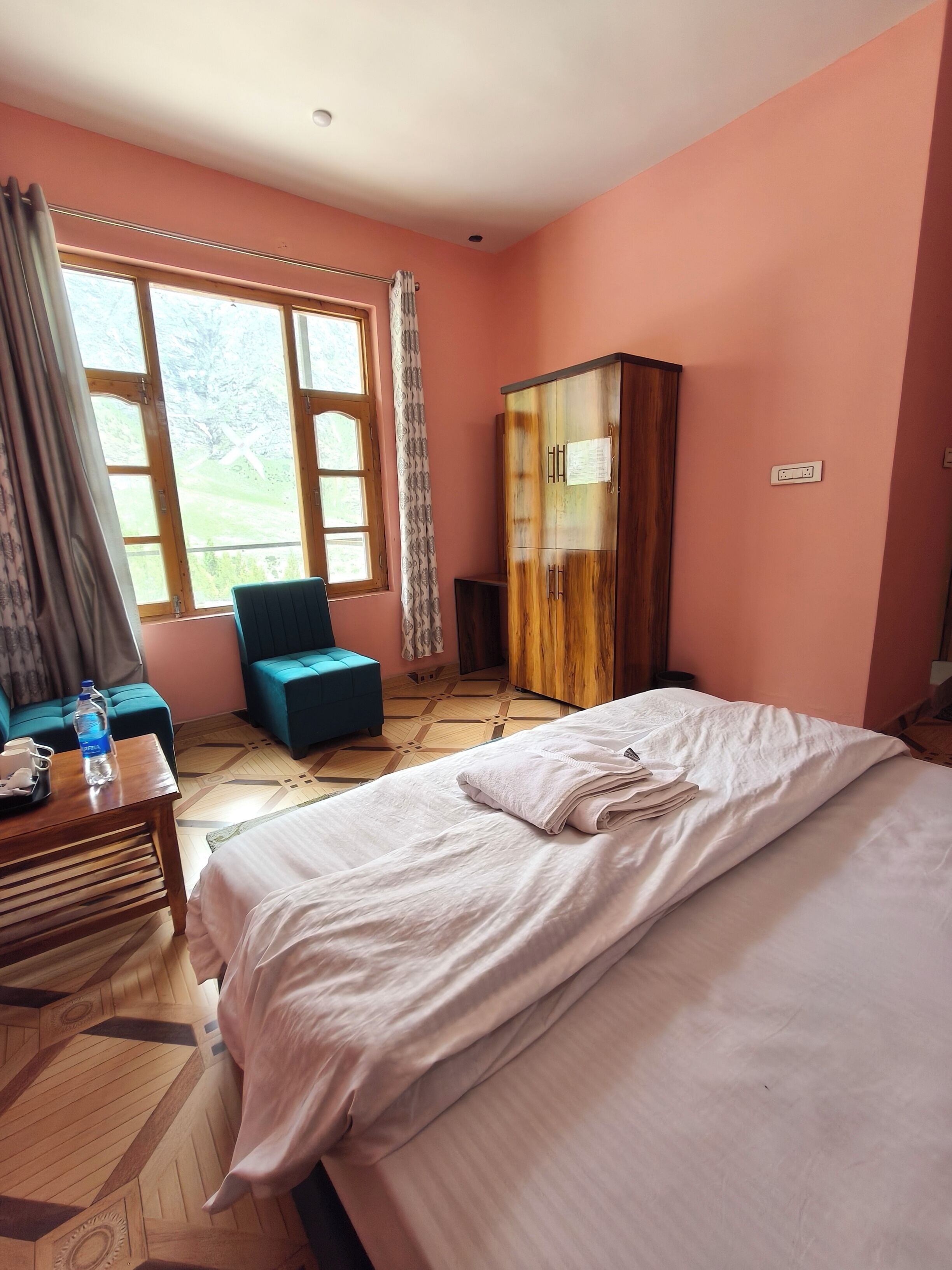 Una habitación de hotel con cama, un sillón azul, un armario de madera y una ventana con cortinas.