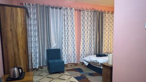 Ein Zimmer mit einem Bett, einem blauen Sessel, einem kleinen Tisch mit Wasserkocher, gemusterten Vorhängen und einem Boden mit geometrischem Muster.