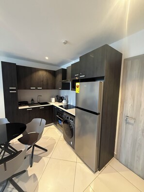 Une cuisine moderne avec des meubles en bois foncé, un réfrigérateur en acier inoxydable, un lave-linge et un coin repas comprenant une table et des chaises noires.