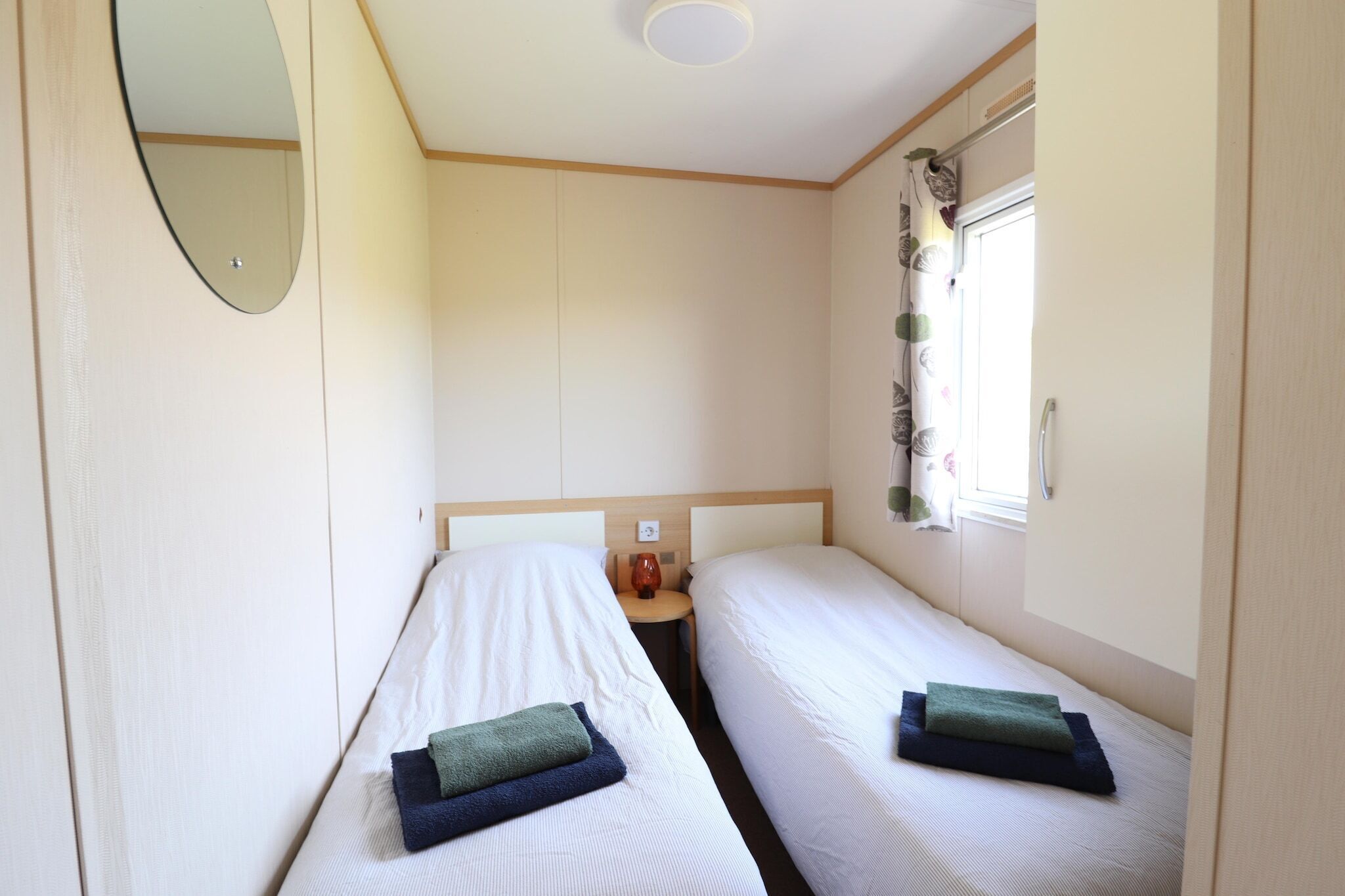 A small, single-room accommodation with two beds, a bedside table, a mirror, and a window with curtains.