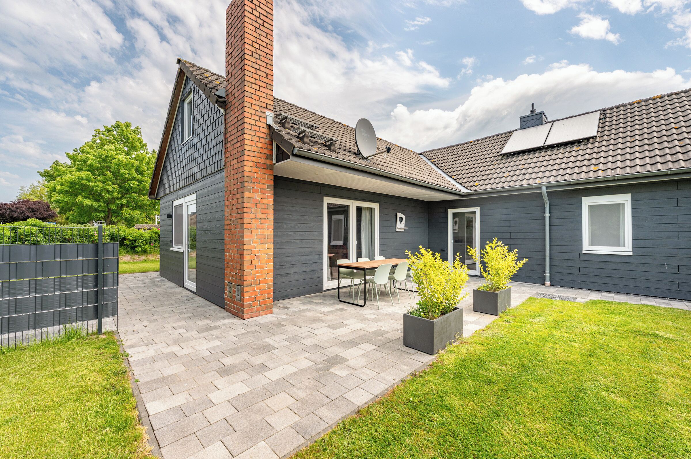 Ein modernes Haus mit Backsteinschornstein, grauer Fassade und einer gepflasterten Terrasse mit Gartenmöbeln.