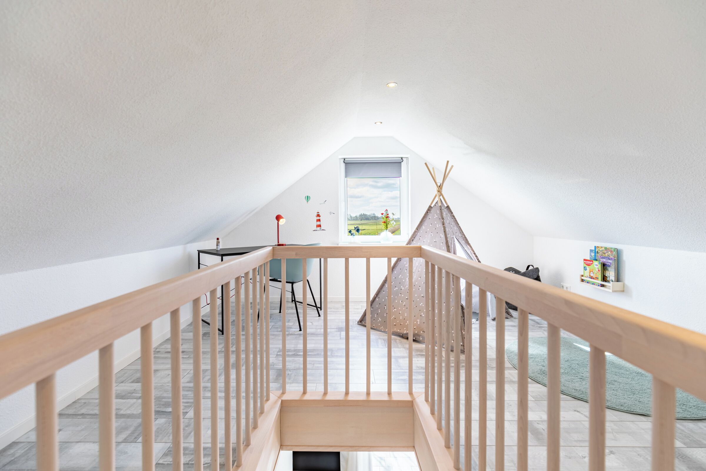 Ein geräumiges Dachzimmer mit einer Holztreppe, einem kleinen Tisch, einem Stuhl, einem Bücherregal und einem Fenster mit Blick ins Freie.