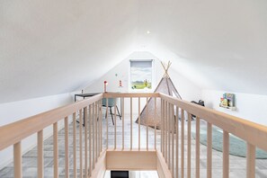 A spacious attic room with a wooden staircase, a small table, a chair, a bookshelf, and a window with a view of the outdoors. - Cottage "Cottage am Deich Whg. (Uelvesbüll)