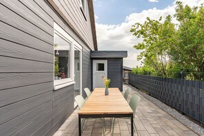 A wooden table with chairs and a vase of flowers in a covered outdoor area with a view of a house and trees. - Cottage "Cottage am Deich Whg. (Uelvesbüll)