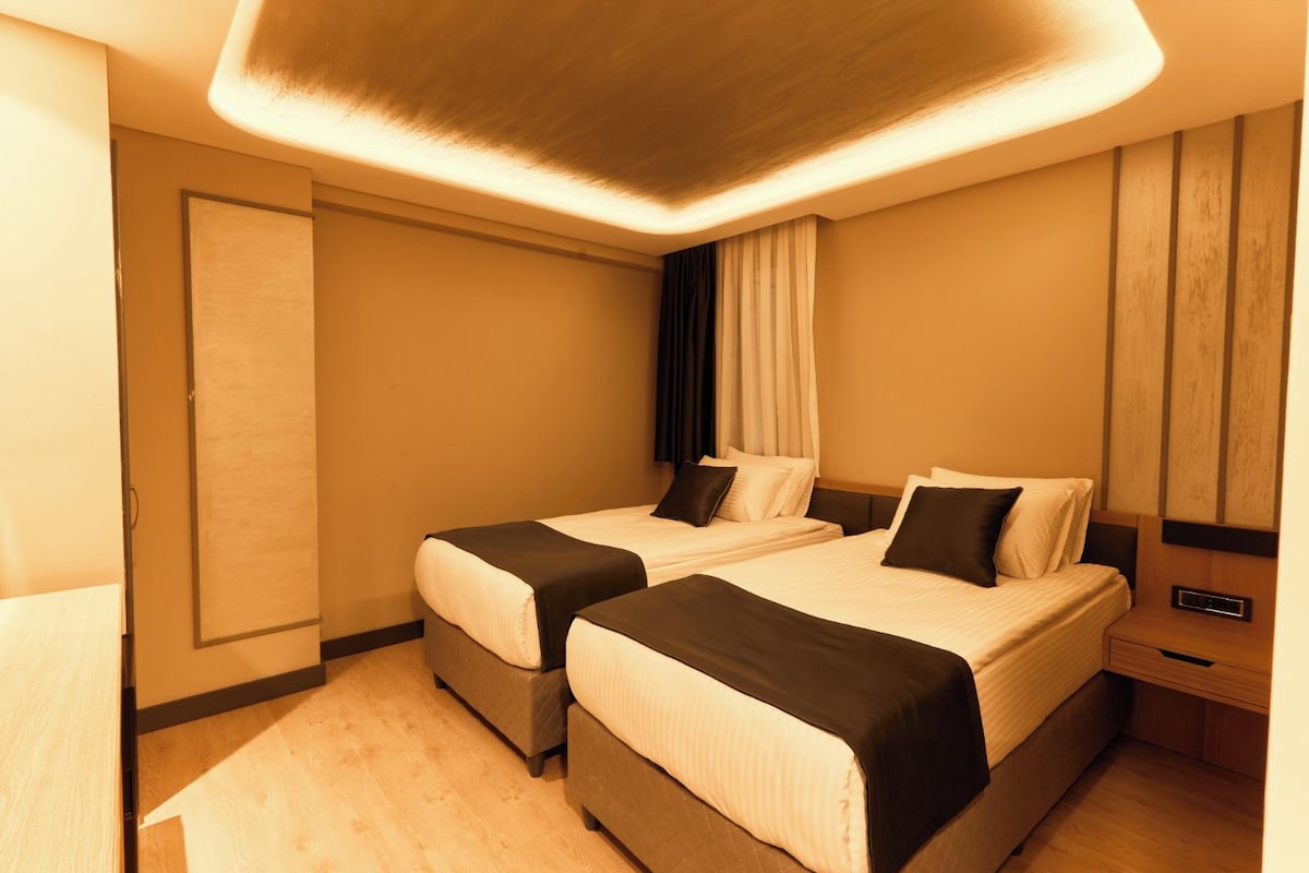 A hotel room with two beds, a wooden floor, a ceiling light fixture, and a nightstand with a lamp.