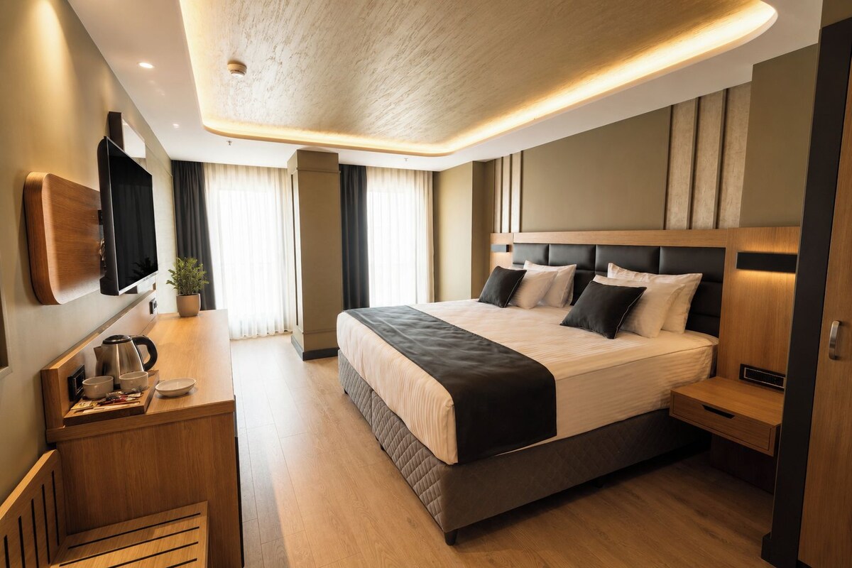 A modern hotel room with a large bed, bedside tables, a flat-screen TV, and a wooden desk with a kettle.