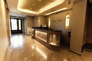 Zona de recepción de hotel con piso de mármol, mostrador de recepción con panel de vidrio y señalización en la pared.