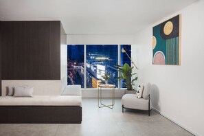 Una sala moderna con un sofá, una mesita auxiliar, una silla, un ventanal con vistas a la ciudad y un cuadro abstracto colgado en la pared.