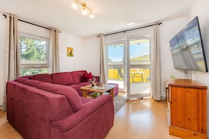Ein Wohnzimmer mit einem roten Sofa, einem Holzschrank, einem Tisch mit Buch und Wein sowie einem Fernseher an der Wand.