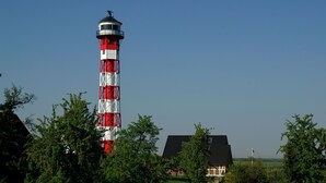 Un faro con un patrón rojo y blanco a rayas, rodeado de follaje y una casa de techo oscuro.