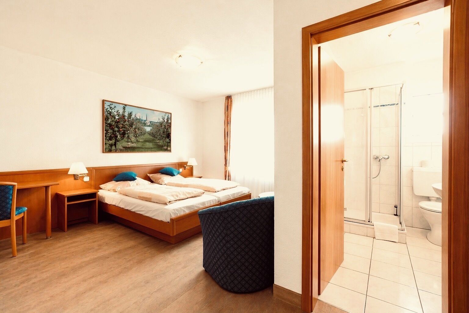 A hotel room with a bed, a desk, a chair, a small table, a painting on the wall, and a shower area.